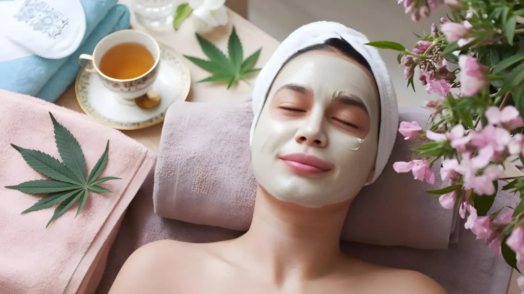 A smiling, relaxed woman lying down with her head on a folded towel. She is wearing a pale-coloured face mask. Beside her is a cup of tea and fresh cannabis leaves.