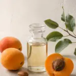 Editorial photograph of a clear glass bottle of pale apricot kernel oil with two whole apricots, one halved apricot, loose kernels and a leafy branch. Above are the words Apricot Kernel Oil: For Skin, Hair & Formulating.