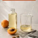 Cosmetic formulation workbench with a clear bottle and glass beaker of pale apricot kernel oil, a stirring rod, mini whisk, digital scale, whole and halved apricots, and loose kernels. Text reads Apricot Kernel Oil: Light, Versatile, Heat Stable, with 420formulator.com below.